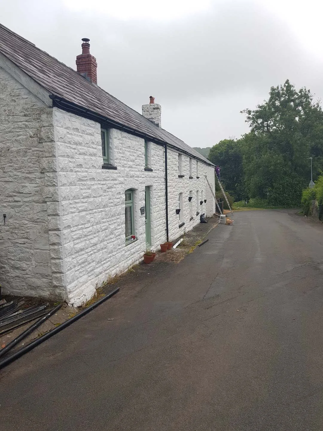 Long white stone cottage with a roofer on a ladder at the eaves and new black gutter lengths on the ground